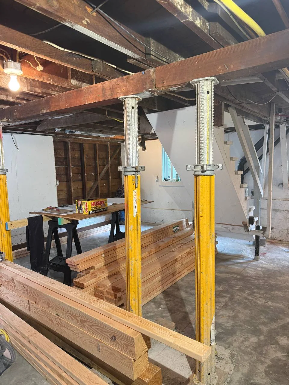 Yellow temporary supports, exposed ceiling framing and stacked dimensional lumber in the basement